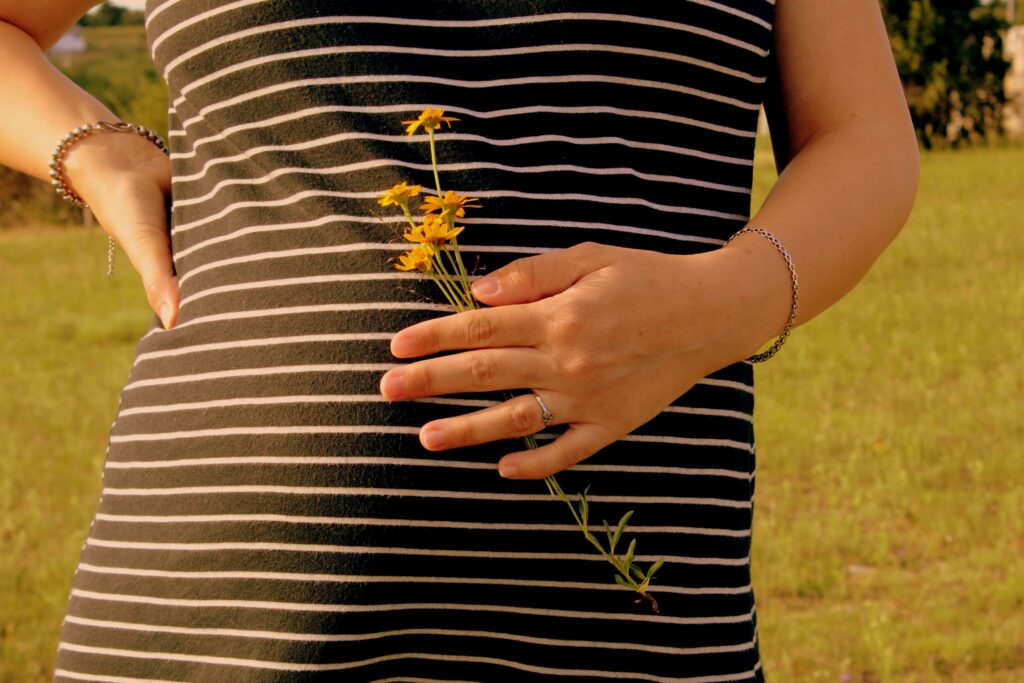 Close-up of a pregnant woman holding yellow wildflowers in a striped dress outdoors.